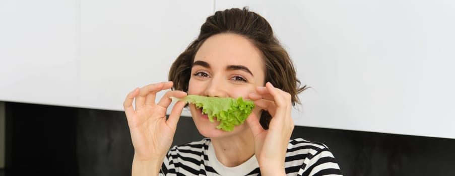 Close up portrait of young woman, vegetarian girl, likes eating vegetables, posing with lettuce leaf and smiling, posing in the kitchen. Concept of healthy food and diet.