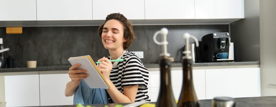 Close up of happy, smiling young woman writing in notebook, creating recipe, list of meals for this week, sitting near vegetables and olive oil, cooking in the kitchen.