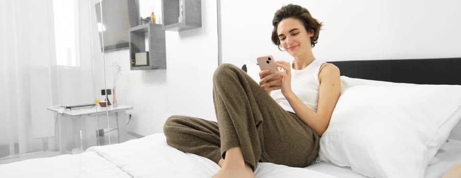 Portrait of beautiful female model using smartphone and sitting in bed, resting in her bedroom, scrolling social media or websites on mobile phone.