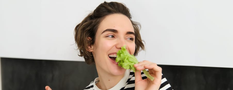 Portrait of carefree vegetarian girl, eating vegies, bite lettuce leaf with happy smiling face, having healthy snack, likes vegetables, stands in the kitchen.