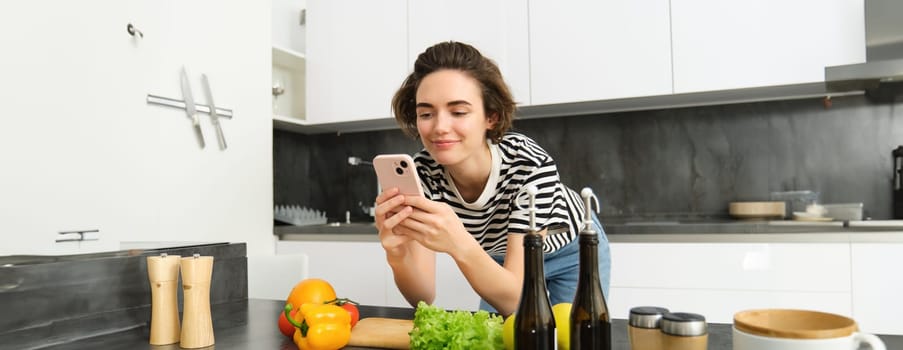 Woman bending on kitchen counter with smartphone, looking for healthy recipe online on mobile phone, standing next to chopping board and vegetables, cooking ingredients.