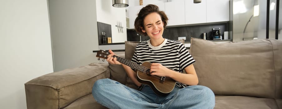 Young woman playing ukulele with passion, singing song, strumming strings, sitting on sofa in living room at home. Lifestyle and music concept