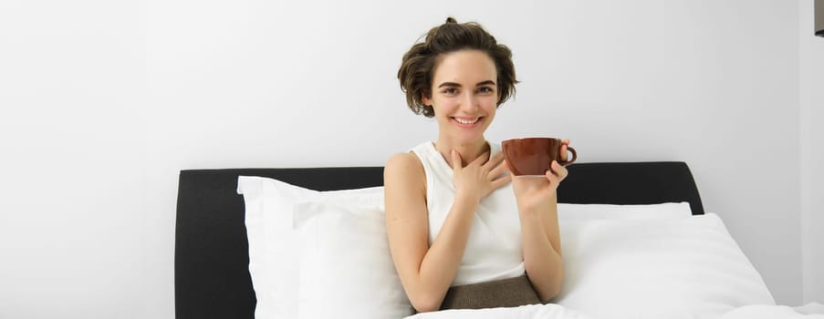 Beautiful young woman with cup of coffee, drinking cappuccino in bed and smiling.