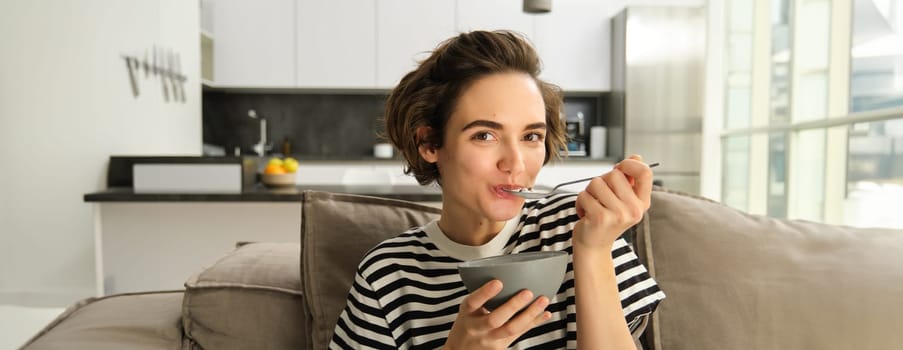 Portrait of young woman eating granola, bowl of cereals with milk, sitting on sofa and having her breakfast.