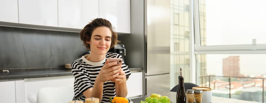 Portrait of cute modern woman cooking in the kitchen, using smartphone, reading recipe, searching meals for breakfast, leaning on counter with vegetables and chopping board.