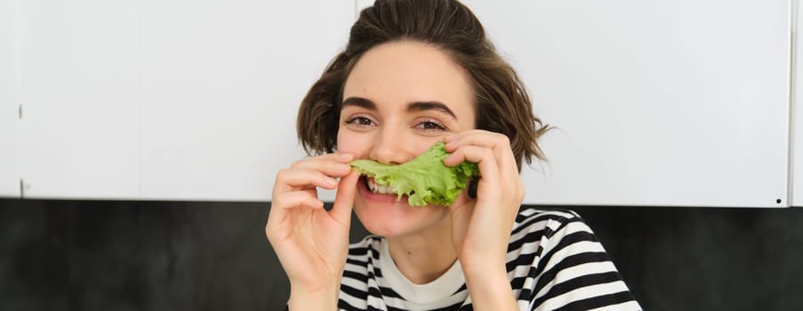 Close up portrait of young woman, vegetarian girl, likes eating vegetables, posing with lettuce leaf and smiling, posing in the kitchen. Concept of healthy food and diet.