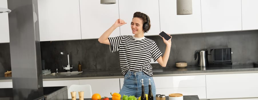 Portrait of happy dancing woman, cooking food in the kitchen, listening music in wireless headphones, chopping vegetables for salad, preparing healthy food and enjoying favourite song.