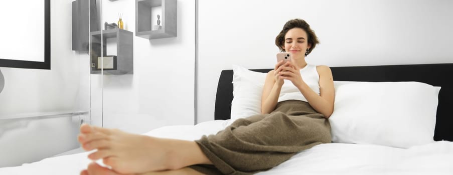Photo of young woman resting in bedroom, lying in bed and using smartphone, looking at mobile phone and smiling, spending relaxing time at home.