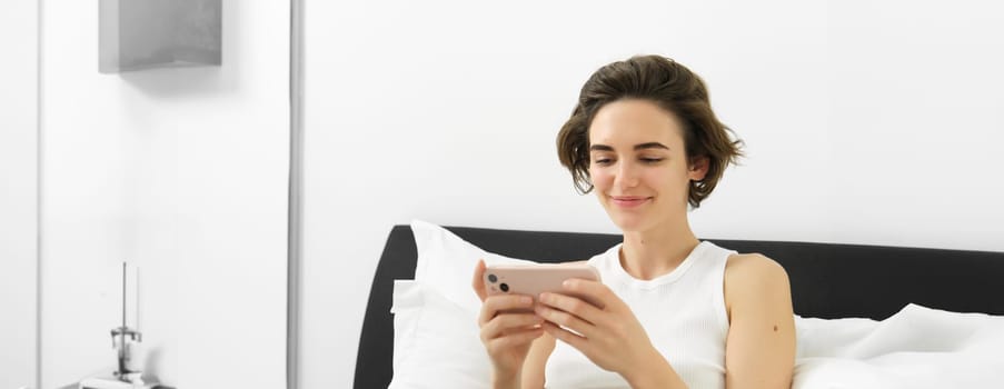Cute young woman with mobile phone in hands, lying in bed in tank top, looking at smartphone with pleased smile.