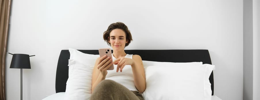 Portrait of young modern woman lying in her bed and using mobile phone, resting in bedroom and looking at smartphone screen.