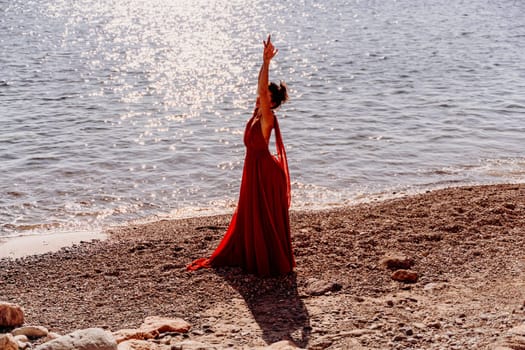 Woman red dress sea. Woman in a long red dress posing on a beach with rocks on sunny day. Girl on the nature on blue sky background