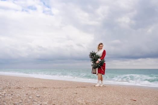 Blond woman holding Christmas tree by the sea. Christmas portrait of a happy woman walking along the beach and holding a Christmas tree in her hands. Dressed in a red coat, white dress