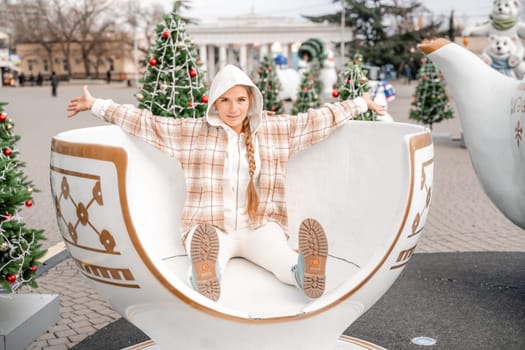 Woman Christmas Square. She sits in a large white cup, dressed in a light suit. With trees decorated with Christmas tinsel in the background.