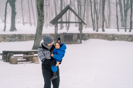 Mom carries a small child in her arms through a snowy forest and looks at his face. High quality photo