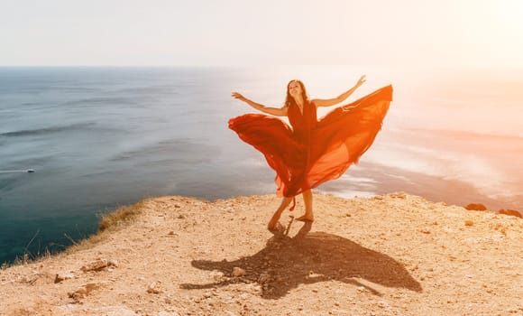 Side view a Young beautiful sensual woman in a red long dress posing on a rock high above the sea during sunrise. Girl on the nature on blue sky background. Fashion photo.