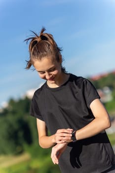 Teenage girl in black clothes checking her fitness watch after a workout. Tired runner girl controlling pulsations checking smartwatch.