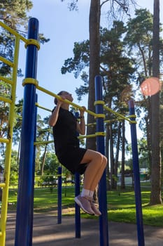 Portrait of beautiful teen age athletic girl training outdoors at open air gym at summer sunny day, gain hands muscles on horizontal bar. fitness, healthy lifestyle.