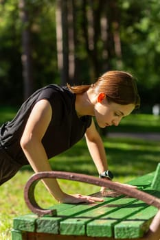 Beautiful young sporty woman in black t-shirt, black shorts and pink trainers warming up exercising triceps and chest dip doing push-ups from bench among trees before running.