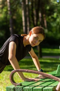 Beautiful young sporty woman in black t-shirt, black shorts and pink trainers warming up exercising triceps and chest dip doing push-ups from bench among trees before running.