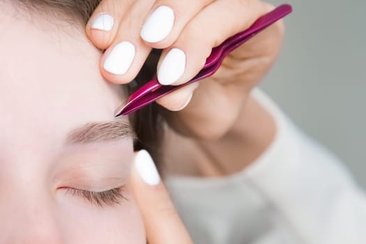 A makeup artist plucks a young woman's eyebrows with tweezers. Beautiful thick eyebrows close-up. Professional makeup and cosmetological skin care in a beauty salon