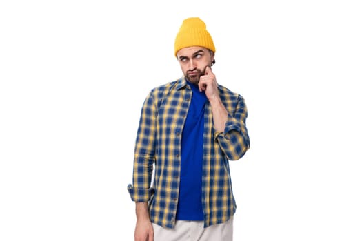 young pensive smart European brunette man with a beard and mustache is dressed in a yellow hat and blue shirt on a white background with copy space.