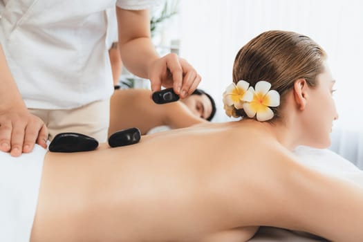 Hot stone massage at spa salon in luxury resort with day light serenity ambient, blissful couple customer enjoying spa basalt stone massage glide over body with soothing warmth. Quiescent