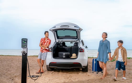 Family vacation trip traveling by the beach with electric car, lovely family taking luggage out while charging EV car battery with clean energy. Alternative family travel by eco-friendly car.Perpetual