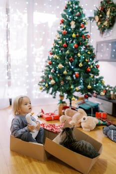 Little girl with a toy duck sits in a gift box with a striped cat near the Christmas tree. High quality photo