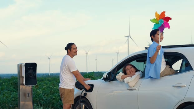 Concept of progressive happy family enjoying their time at wind farm with electric vehicle. Electric vehicle driven by clean renewable energy from wind turbine generator for charging station.