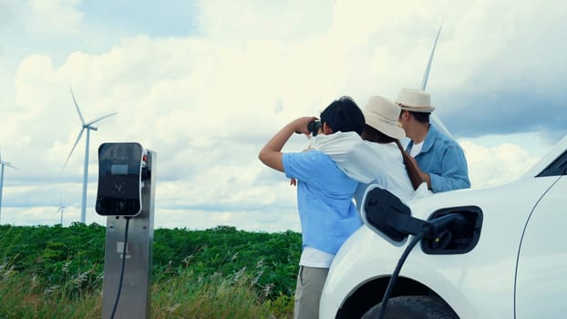 Concept of progressive happy family enjoying their time at wind farm with electric vehicle. Electric vehicle driven by clean renewable energy from wind turbine generator for charging station.