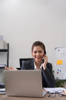 Cheerful young asian business woman talking on phone working in office. Happy positive Asian businesswoman company manager wearing suit making call on cellphone sitting at workplace.