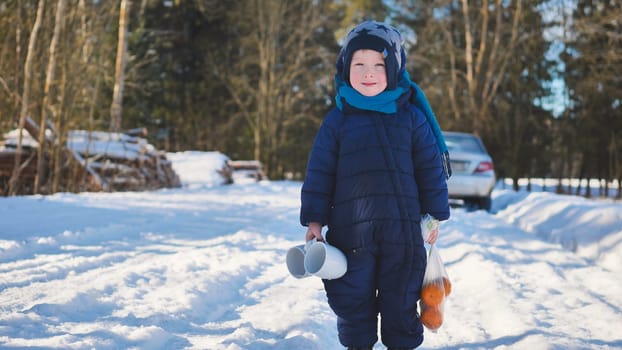 A boy walks down a snowy road on a sunny winter day with mugs and tangerines