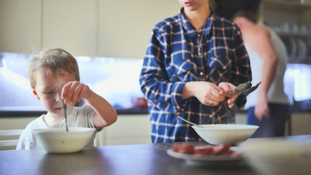 The child sits at the table in front of the bowl. The parents bring him a dish