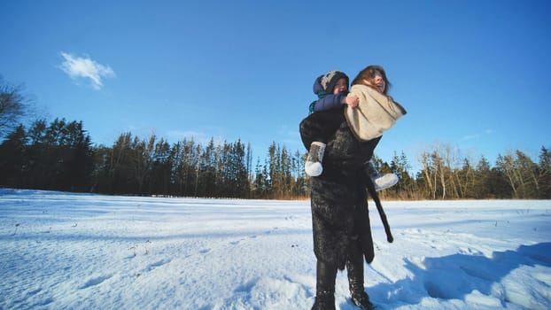 A mother carries her son across a field on her shoulders in winter