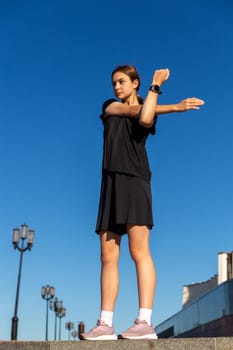 Young, fit and sporty girl in black clothes stretching after the workout in the urban city park. Fitness, sport, urban jogging and healthy lifestyle concept.