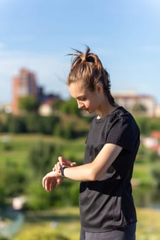 Teenage girl in black clothes checking her fitness watch after a workout. Tired runner girl controlling pulsations checking smartwatch.