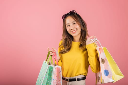 In this studio shot on a pink background, a cute and happy Asian woman, sporting sunglasses, stands with delight, clutching multicolored shopping bags. She's a model of joyful shopping.