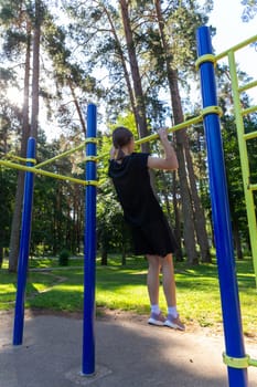 Portrait of beautiful teen age athletic girl training outdoors at open air gym at summer sunny day, gain hands muscles on horizontal bar. fitness, healthy lifestyle.