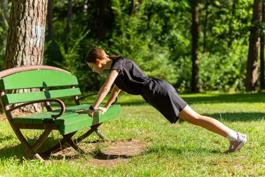 Beautiful young sporty woman in black t-shirt, black shorts and pink trainers warming up exercising triceps and chest dip doing push-ups from bench among trees before running.