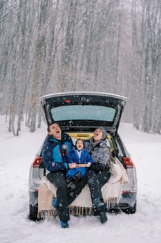 Parents and a small child catching snowflakes in their mouths while sitting in the trunk of a car under snowfall in the forest. High quality photo