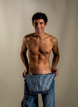 A shirtless man holding a pair of jeans overalls, naked underneath, smiling to the camera, undressing in a sexy manner
