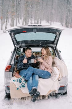 Woman laughing with cup sitting next to man sipping coffee and holding thumb up in car trunk. High quality photo