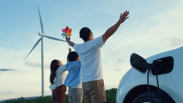 Concept of progressive happy family enjoying their time at wind farm with electric vehicle. Electric vehicle driven by clean renewable energy from wind turbine generator for charging station.