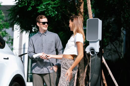 Young couple travel with EV electric car charging in green sustainable city outdoor garden in summer shows urban sustainability lifestyle by green clean rechargeable energy of electric vehicle innards