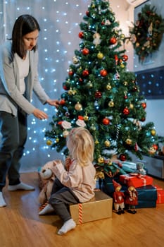 Little girl sits on gifts and watches her mother decorate the Christmas tree. Back view. High quality photo