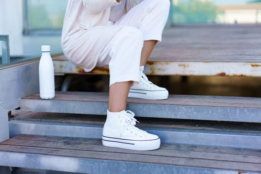 Unrecognizable woman taken a coffee break, sitting on steps near her office building with an eco-friendly ecological metal water bottle. Businesswoman using smartphone.