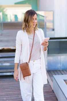 Stylish female browsing social media on cellphone while walking on path outside modern building on sunny day in city