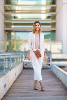 Full body female in stylish outfit with bag and cellphone standing on footbridge outside contemporary building and looking at camera with smile