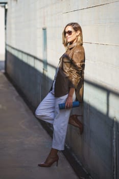 Full body woman in stylish outfit and sunglasses with thermos leaning on sunlit building wall in daytime on city street