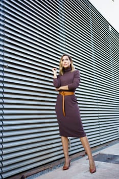 Full body self assured businesswoman in stylish dress with belt and high heeled shoes looking away while standing outside modern building on city street